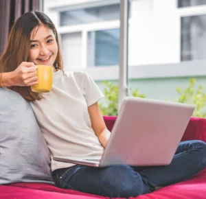 A smiling young woman sitting comfortably on a pink sofa with a laptop on her lap, holding a yellow coffee mug in a bright living room.