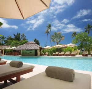 Luxury tropical resort swimming pool with lounge chairs, rolled towels, a poolside bar under a thatched roof, and palm trees under a bright blue sky.